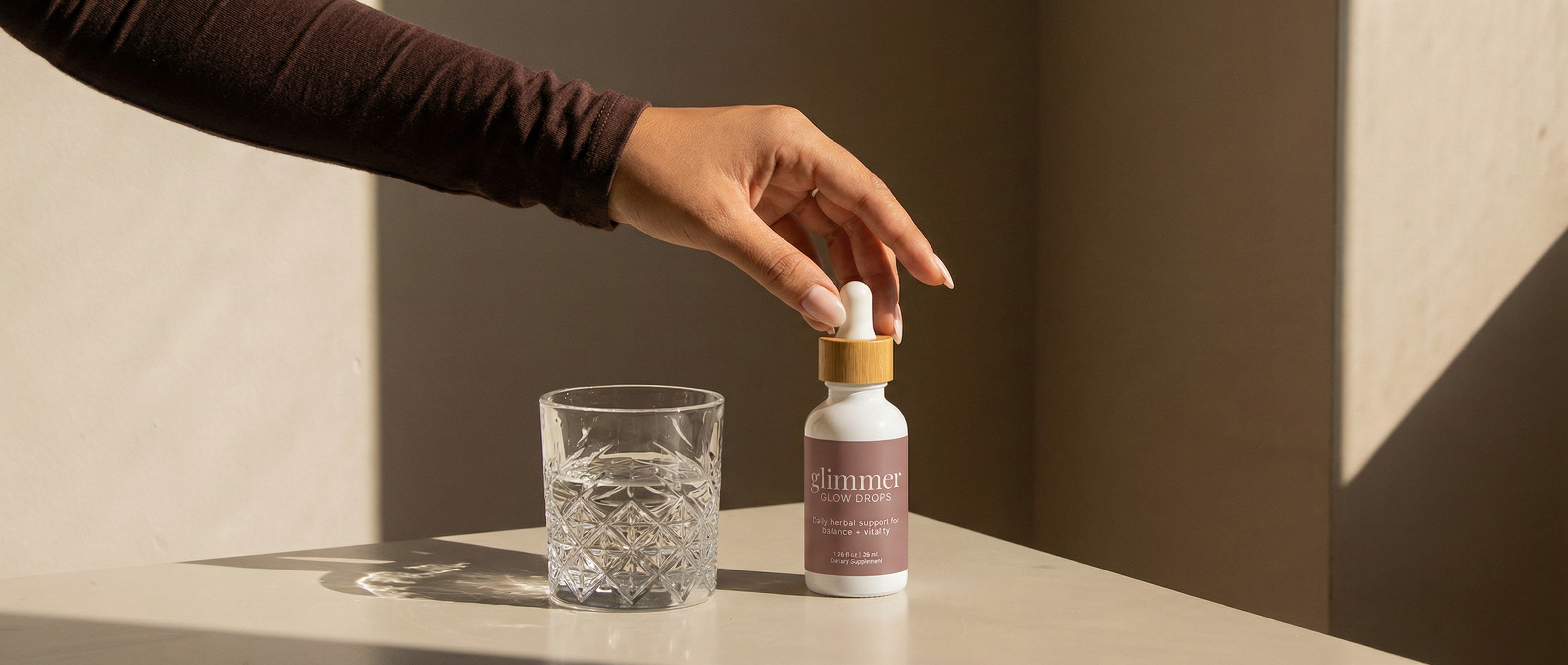 Hand reaching for a Glow Drop bottle with a bamboo cap and pink label next to a glass of water against a neutral background