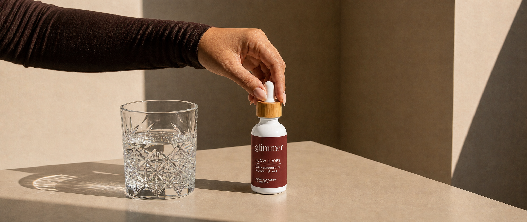 Hand holding a dropper bottle labeled 'Glimmer' over a glass of water on a neutral background